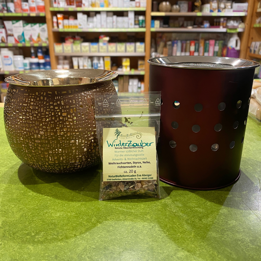 Auf einem Tisch stehen ein bronzefarbener Topf mit Deckel, ein roter perforierter Zylinder und eine kleine Packung Winterzauber-Räucherwerk.