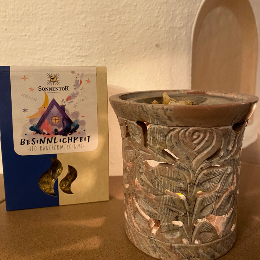 Ein beigefarbener Keramik-Kerzenhalter mit kunstvollen Blumenmustern steht auf einem Holztisch neben einer Packung Sonnentor Bio-Rauchermischung.