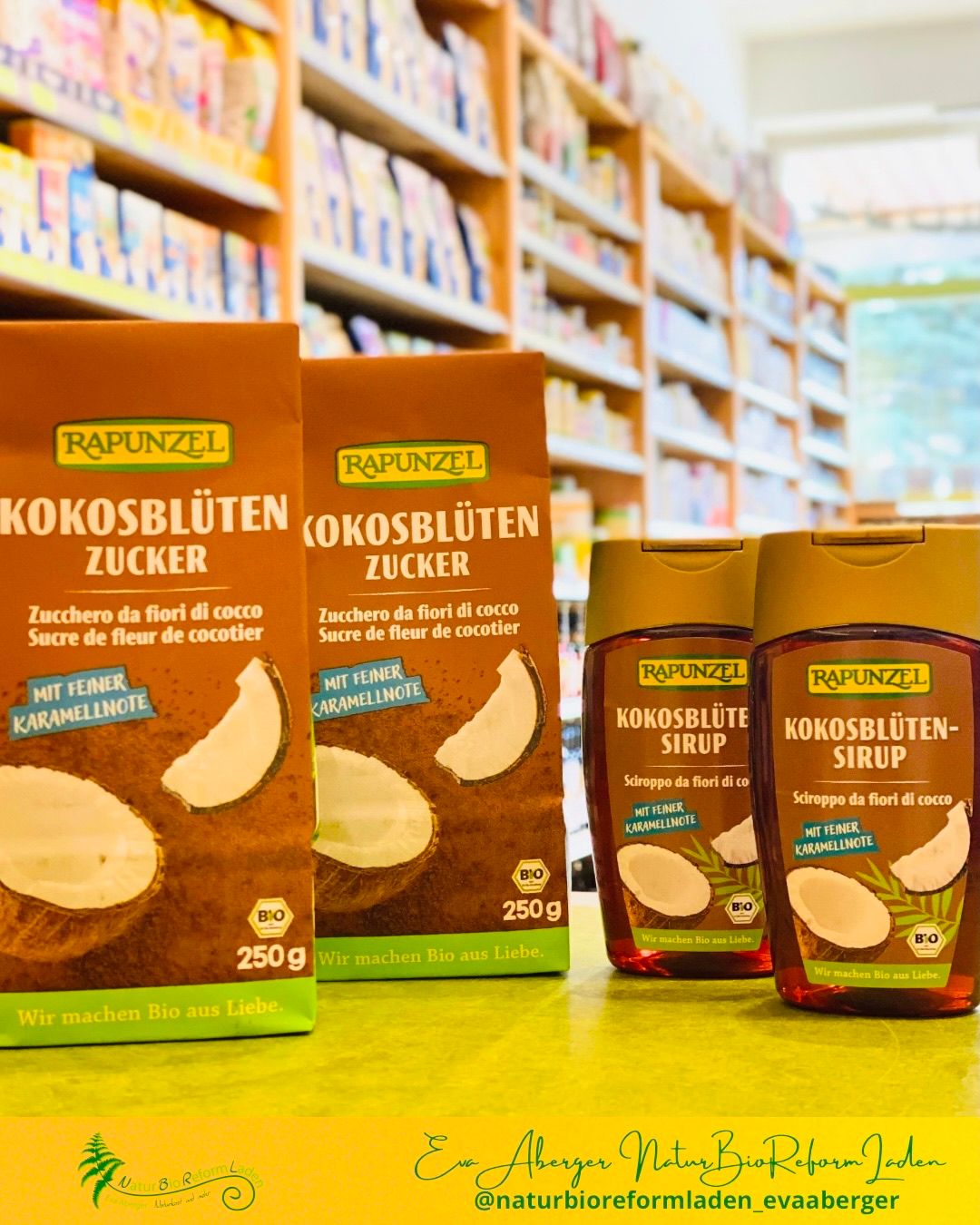 Drei Packungen Rapunzel Kokosblütenzucker sind in einem Geschäft ausgestellt. Jede Packung ist mit 'Bio' und '250g' gekennzeichnet. Zwei der Packungen sind in Pulverform, während die dritte ein Sirup ist.