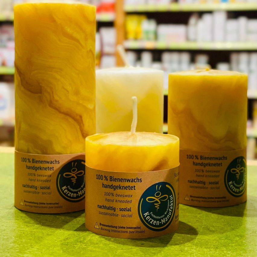 Drei gelbe Kerzen sind auf einem grünen Tisch vor einem Regal mit verschiedenen Produkten ausgestellt. Die Kerzen bestehen zu 100% aus Bienenwachs und sind als nachhaltig beschafft gekennzeichnet.