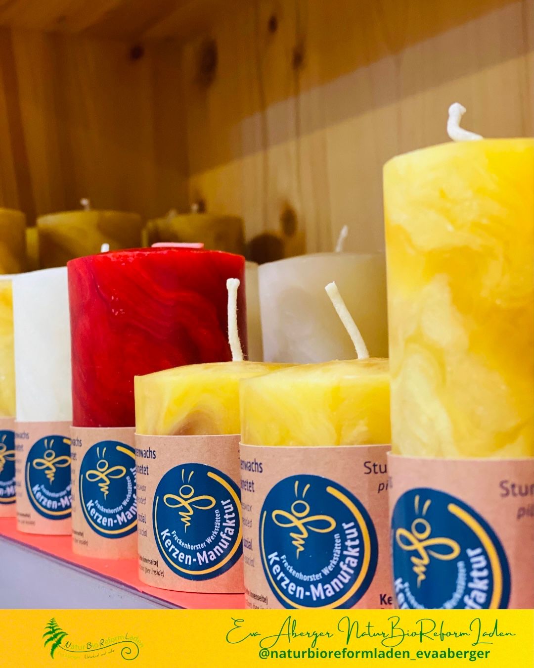 Eine Reihe zylindrischer Kerzen in verschiedenen Farben, einschließlich Gelb, Rot und Weiß, ist auf einem Regal mit hölzerne Hintergrund ausgestellt. Jede Kerze hat einen weißen Docht und ein Etikett.