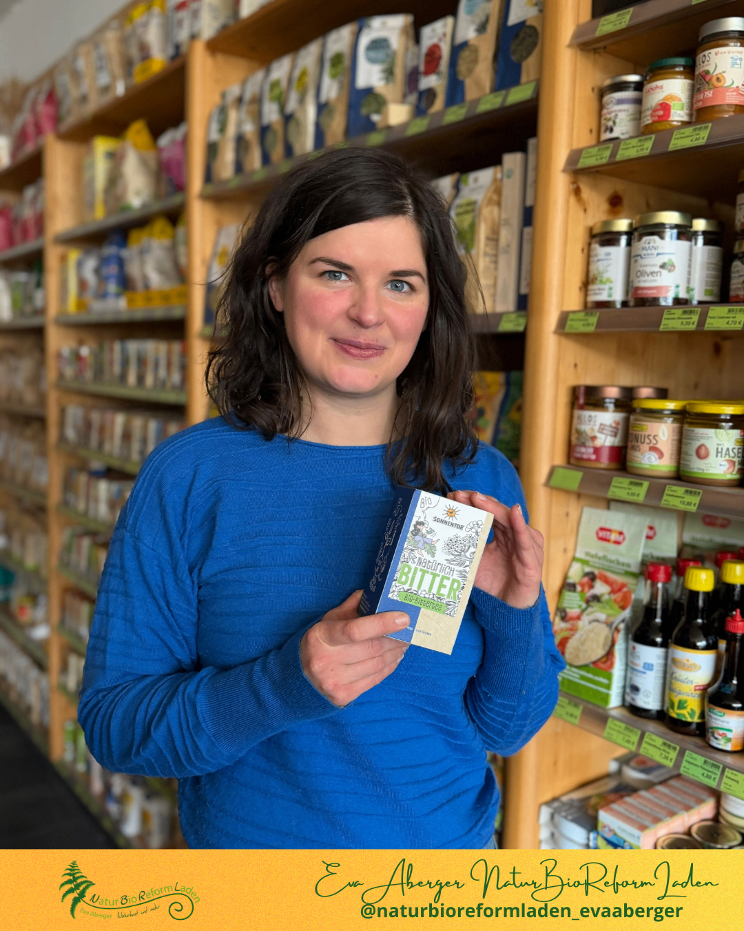 Eine Frau in einem blauen Shirt hält eine Box 'Wadlinich Bitter' vor einem Laden voller verschiedener Produkte.