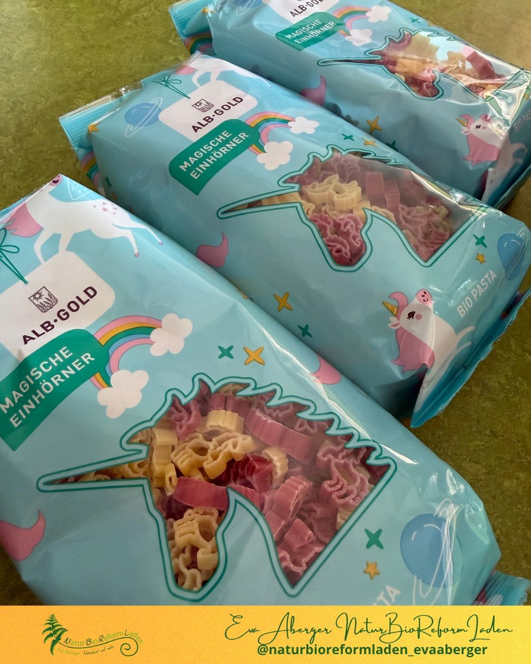 Three bags of Alb-Gold pasta with unicorn and rainbow designs, displayed on a green surface. The pasta is colorful and shaped like unicorns and stars.