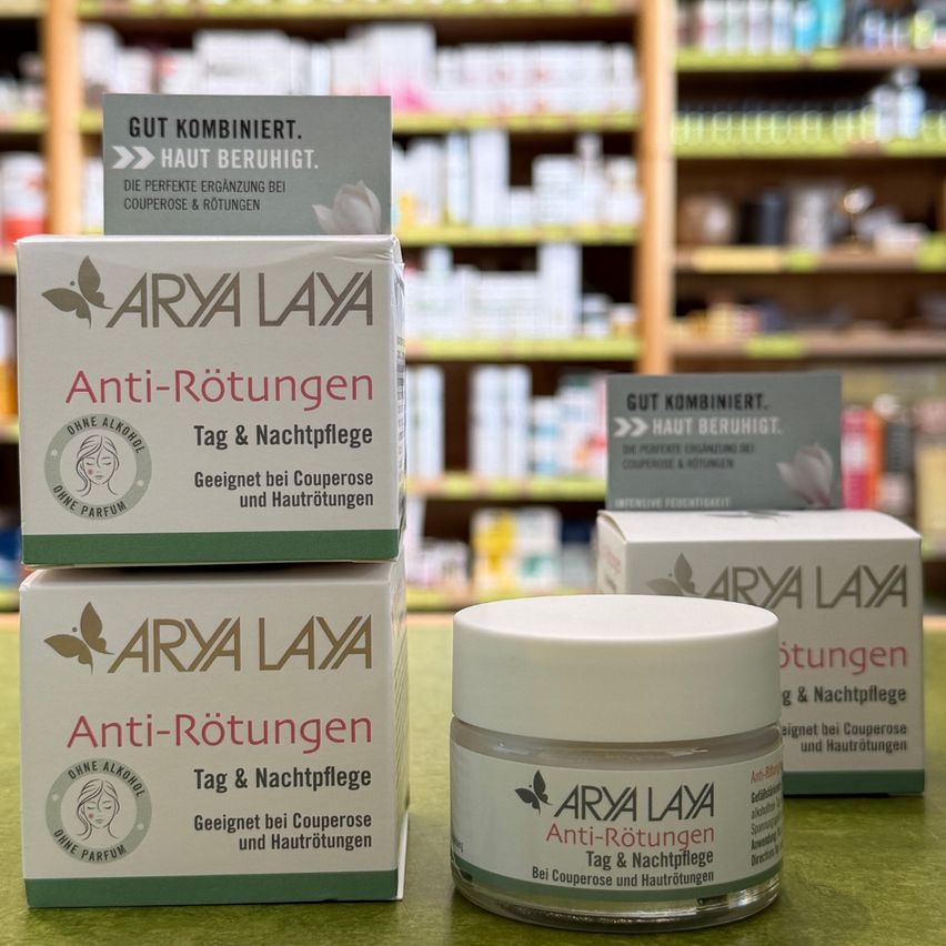 Drei Schachteln Arya Laya Anti-Rötungen Creme und ein Tiegel des gleichen Produkts sind auf einer grünen Oberfläche ausgestellt. Die Schachteln sind übereinander gestapelt und der Tiegel ist davor platziert. Der Hintergrund ist ein verschwommenes Bild einer Apotheke mit Regalen voller verschiedener Produkte.