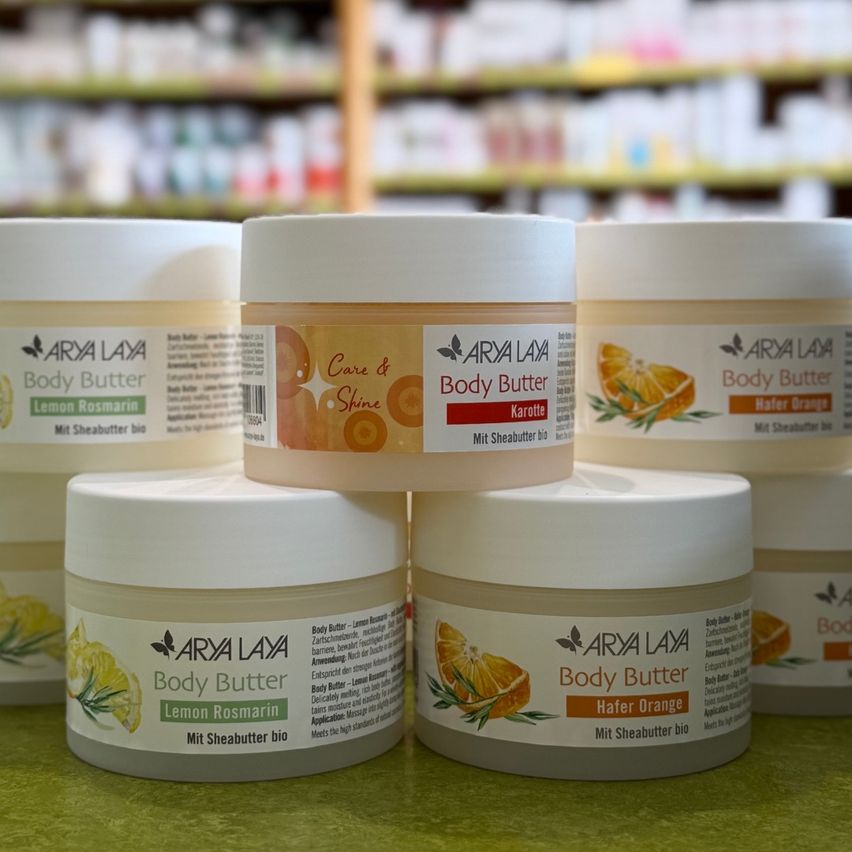 Fünf Behälter mit Arya Lay Körperbuttern mit Zitrone, Rosmarin und Orangenduft stehen auf einer grünen Oberfläche vor einem Ladenregal.
