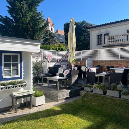 Ein Hinterhof mit Sitzplätzen im Freien hat Tische, Stühle und Pflanzen. Auf einem Tisch steht ein geschlossener Regenschirm. Dahinter steht ein weißes Gebäude mit Balkon.