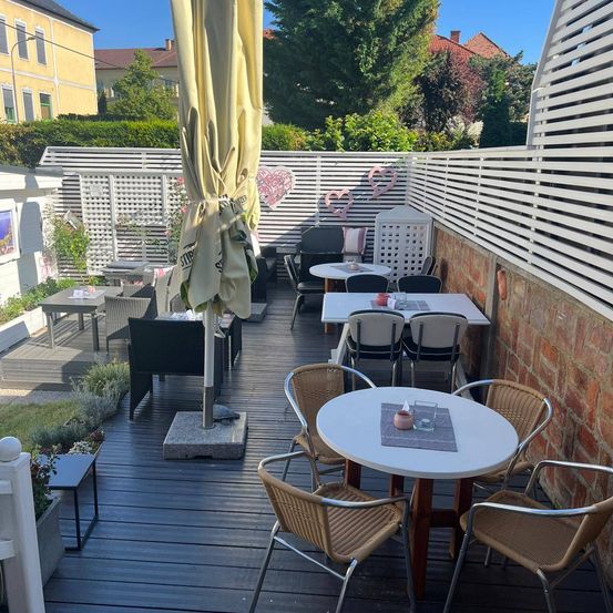 Ein Terrassenbereich zum Essen mit Holzboden, weißen Tischen und Korbstühlen. Ein weißer Regenschirm steht in der Mitte. Der Bereich ist mit weißen Zäunen eingezäunt, und Herzdesigns sind ausgestellt.