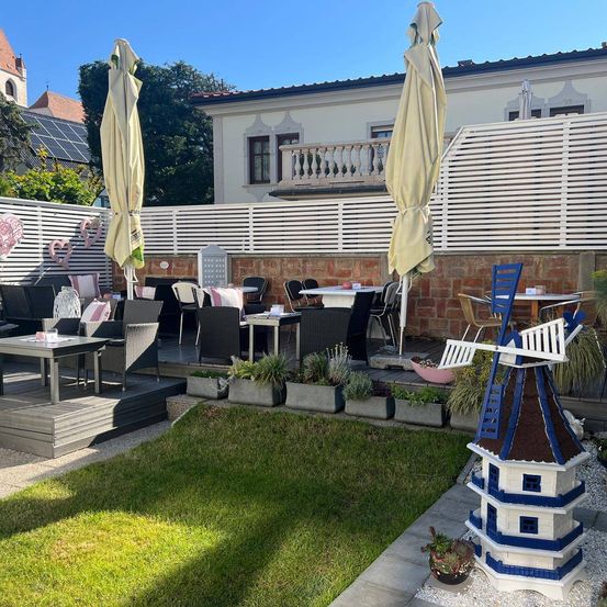 Eine Terrasse mit Tischen, Stühlen und Sonnenschirmen, mit einer Windmühle und Pflanzen. Im Hintergrund befindet sich ein Backsteingebäude mit Balkon.