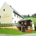Ein gelbes und weißes Gebäude mit einem Balkon und einem Schild mit der Aufschrift 'Stori's Mostachanke' ist von einem grünen Rasen und einem Zaun umgeben. Bäume und ein Berg sind im Hintergrund.