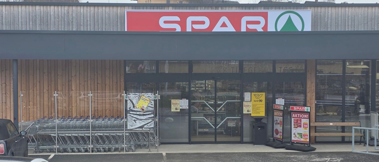 Der Eingang eines Spar-Supermarkts zeigt ein rotes Schild mit weißen Buchstaben, Glastüren und einen Mülleimer. Leere Einkaufswagen sind draußen aufgestellt, und es gibt Werbeschilder für Aktionen.