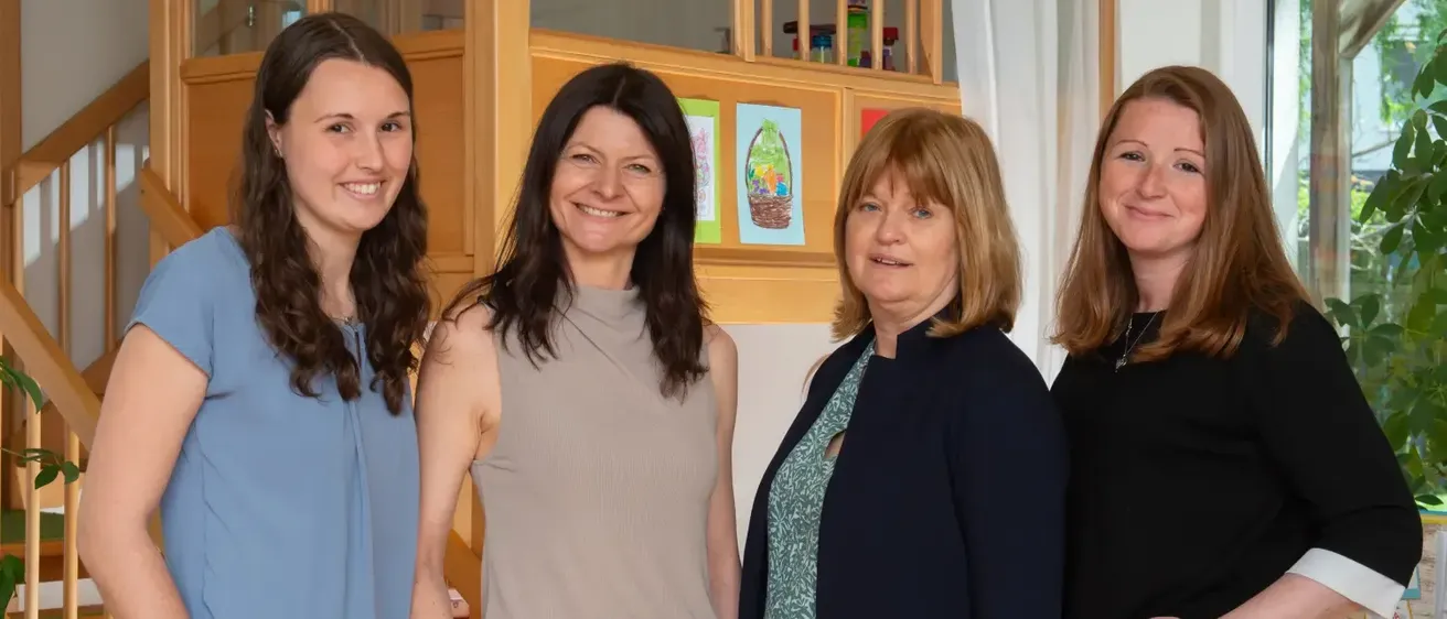 Vier Frauen, die lächeln und für ein Foto posieren, stehen zusammen in einem Raum mit einer Holztreppe und einer weißen Wand.