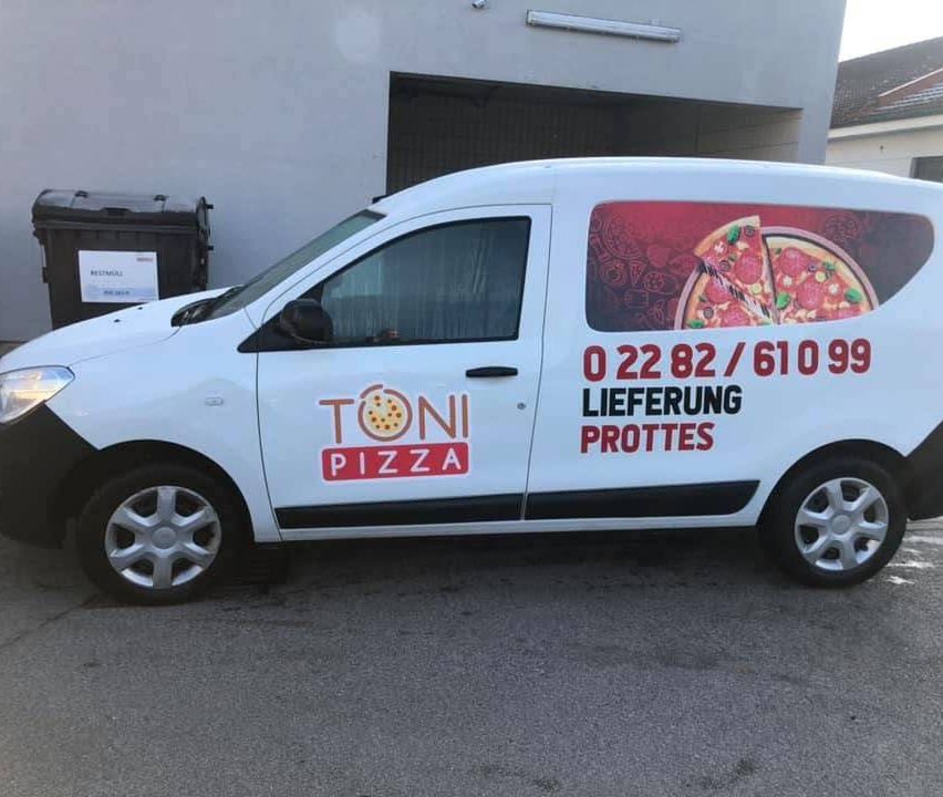 Ein weißer Lieferwagen mit Toni Pizza Logo steht vor einem Gebäude. Der Lieferwagen zeigt eine Pizza-Werbung und eine Liefernummer.