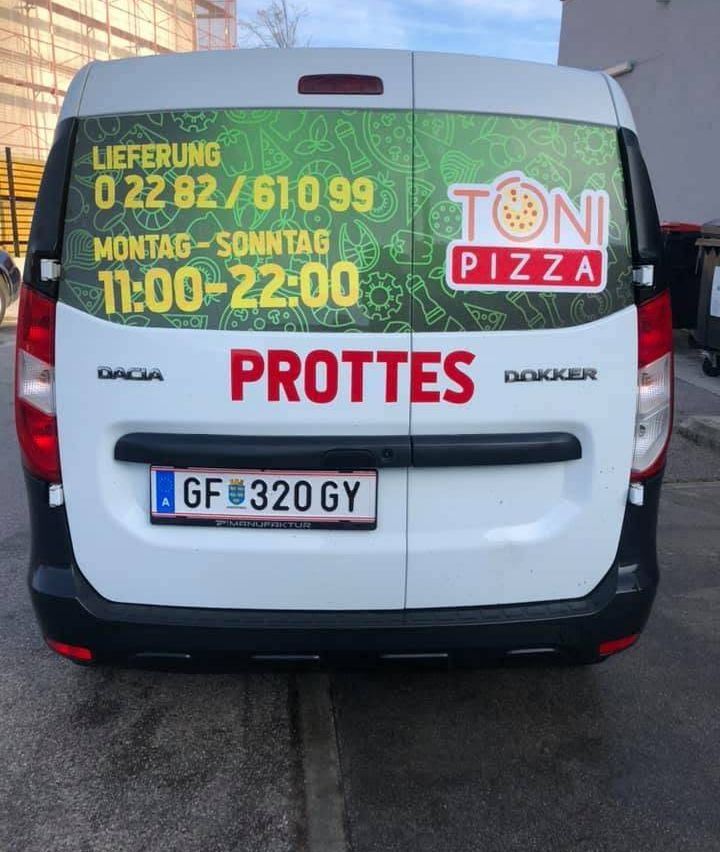 Die Rückseite eines weißen Lieferwagens mit dem Logo 'Dacia', einem Kennzeichen 'GF 320 GY' und dem Wort 'PROTTES'. Es hat einen grün-gelben Aufkleber mit einem Pizzadesign und dem Text 'TONI PIZZA'. Der Lieferwagen zeigt auch die Nummern '0 22 82/610 99' und '11:00-22:00'.