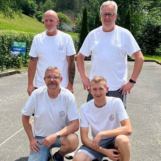 Vier Männer in weißen T-Shirts posieren an einem sonnigen Tag auf einem Parkplatz mit Grünpflanzen im Hintergrund für ein Foto.