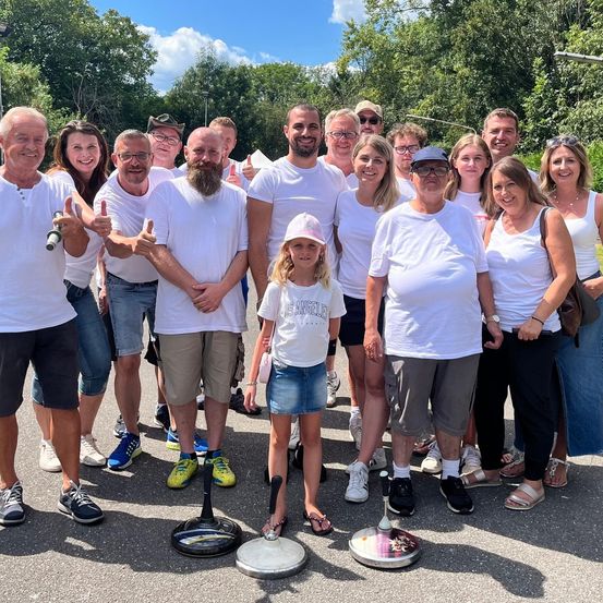 Eine Gruppe von Menschen, einige in weißen T-Shirts, stehen in einer Reihe, lächeln und geben Daumen hoch in einem Park.