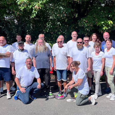 Eine Gruppe Erwachsener, einige mit Bart, in weißen T-Shirts und Turnschuhen, posieren für ein Foto mit Bäumen im Hintergrund.