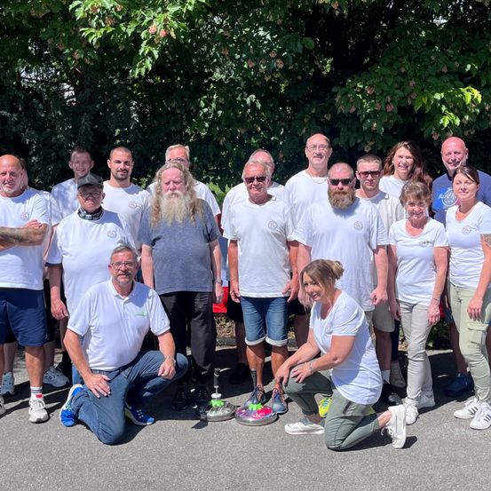 Eine Gruppe Erwachsener, einige mit Bart, in weißen T-Shirts und Turnschuhen, posieren für ein Foto mit Bäumen im Hintergrund.