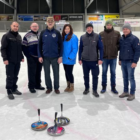Eine Gruppe von sieben Personen steht auf einer Eisbahn, gekleidet in Winterkleidung. Sie scheinen für ein Foto zu posieren.