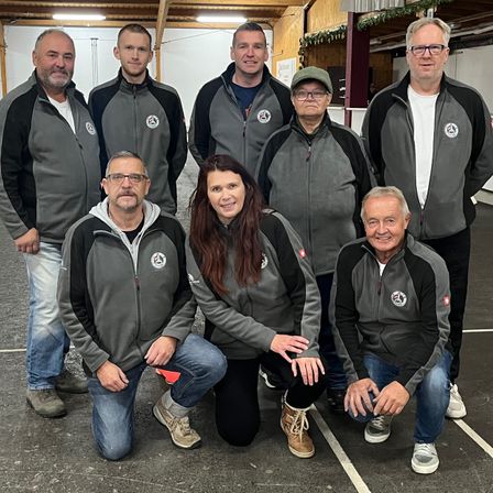 Eine Gruppe von Menschen in passenden grauen Jacken posiert für ein Foto in einer Turnhalle. Sie lächeln und stehen in einer Reihe mit einer Frau, die vorne kniet.