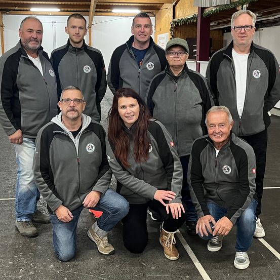 Eine Gruppe von Menschen in passenden grauen Jacken posiert für ein Foto in einer Turnhalle. Sie lächeln und stehen in einer Reihe mit einer Frau, die vorne kniet.