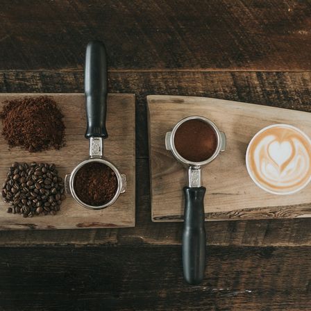 Zwei Kaffee-Utensilien, Kaffeebohnen und ein Latte mit Herzdesign auf einer hölzernen Oberfläche.