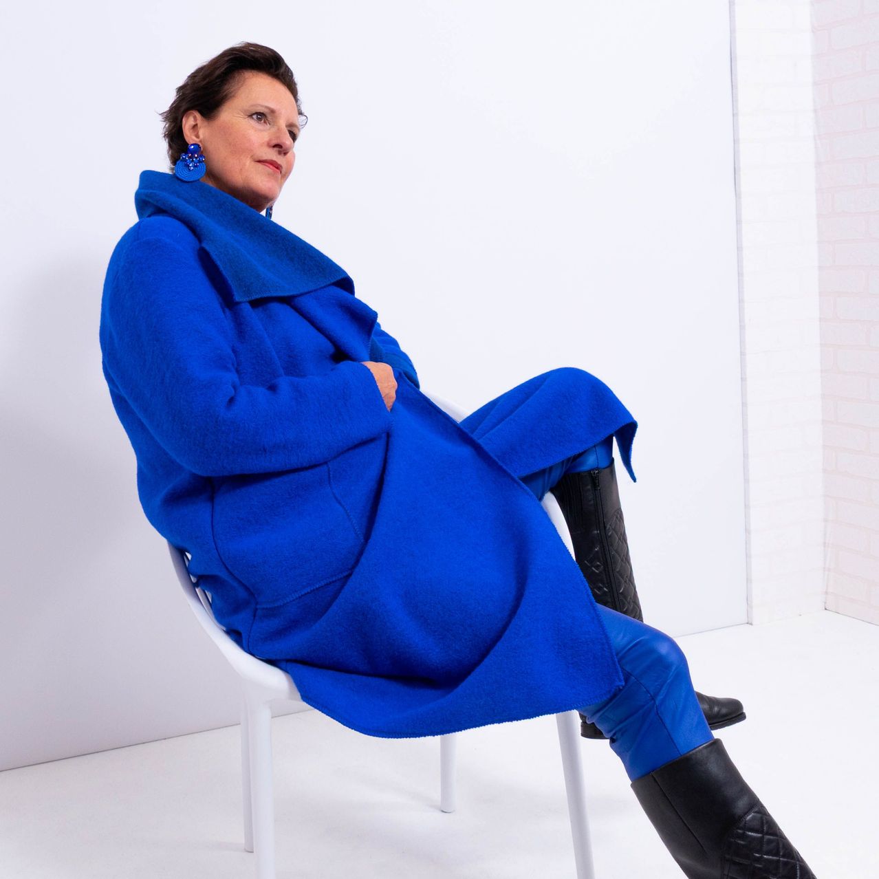 A woman wearing a blue coat and black boots is sitting on a white chair. She is looking sideways with a serious expression.