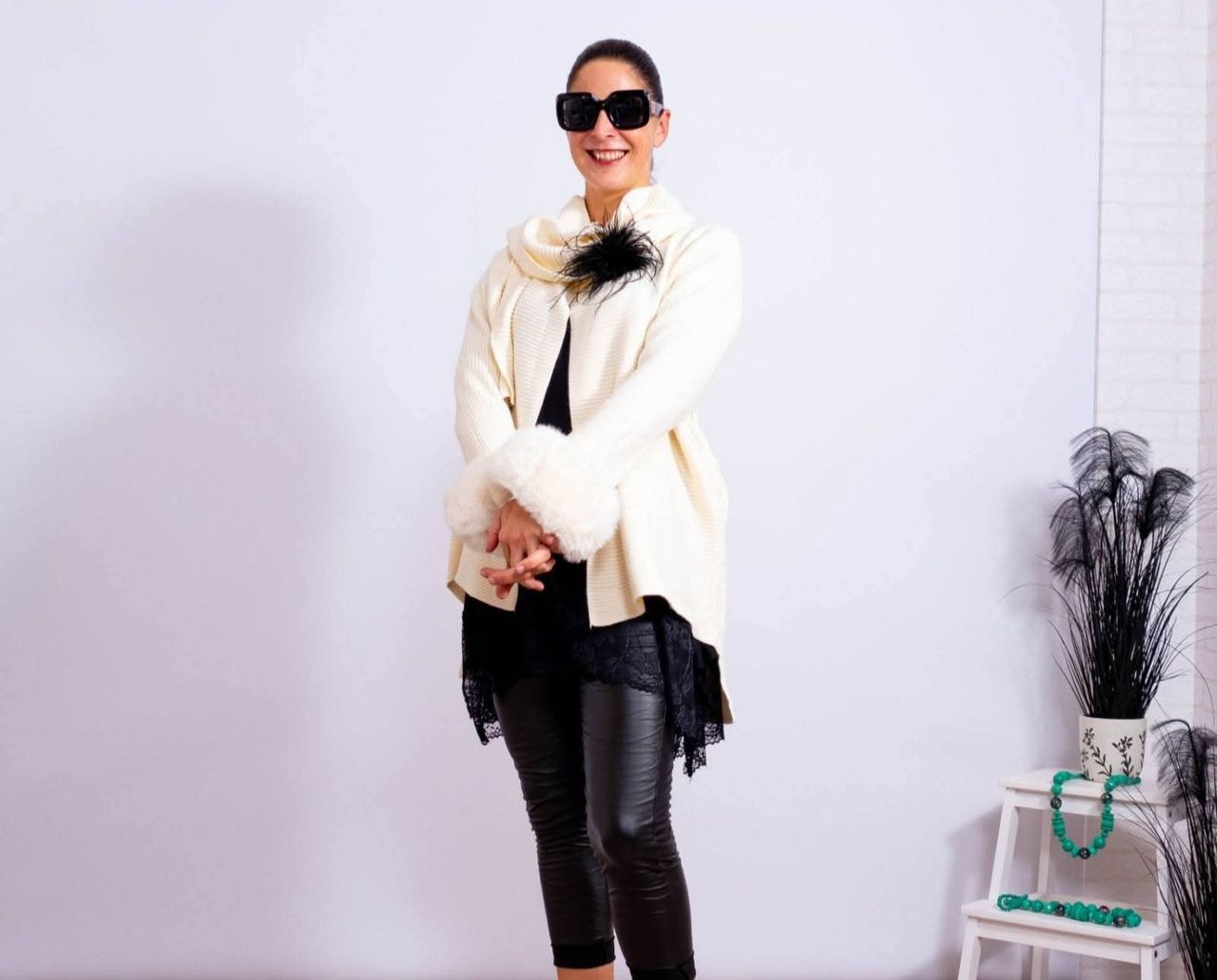 A woman is standing in a room, smiling and posing for a photo. She wears a white fur-trimmed jacket, black lace top, black leather pants, and sunglasses.