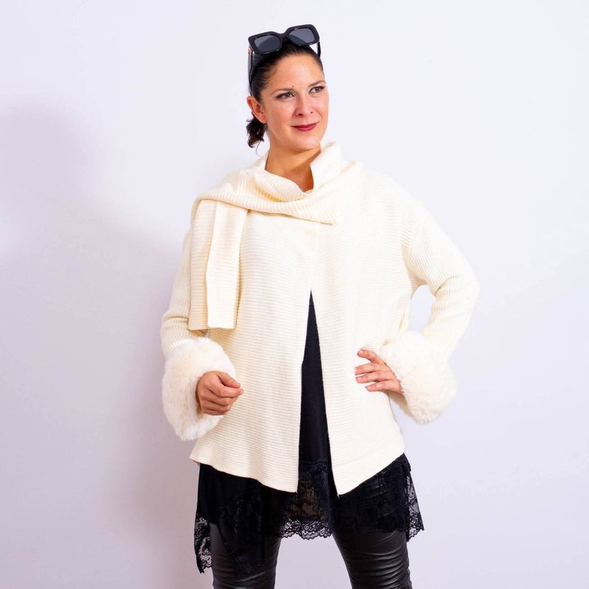 A woman wearing a white, knitted turtleneck sweater with fur sleeves and a black lace skirt, posing for a photo against a plain white background.