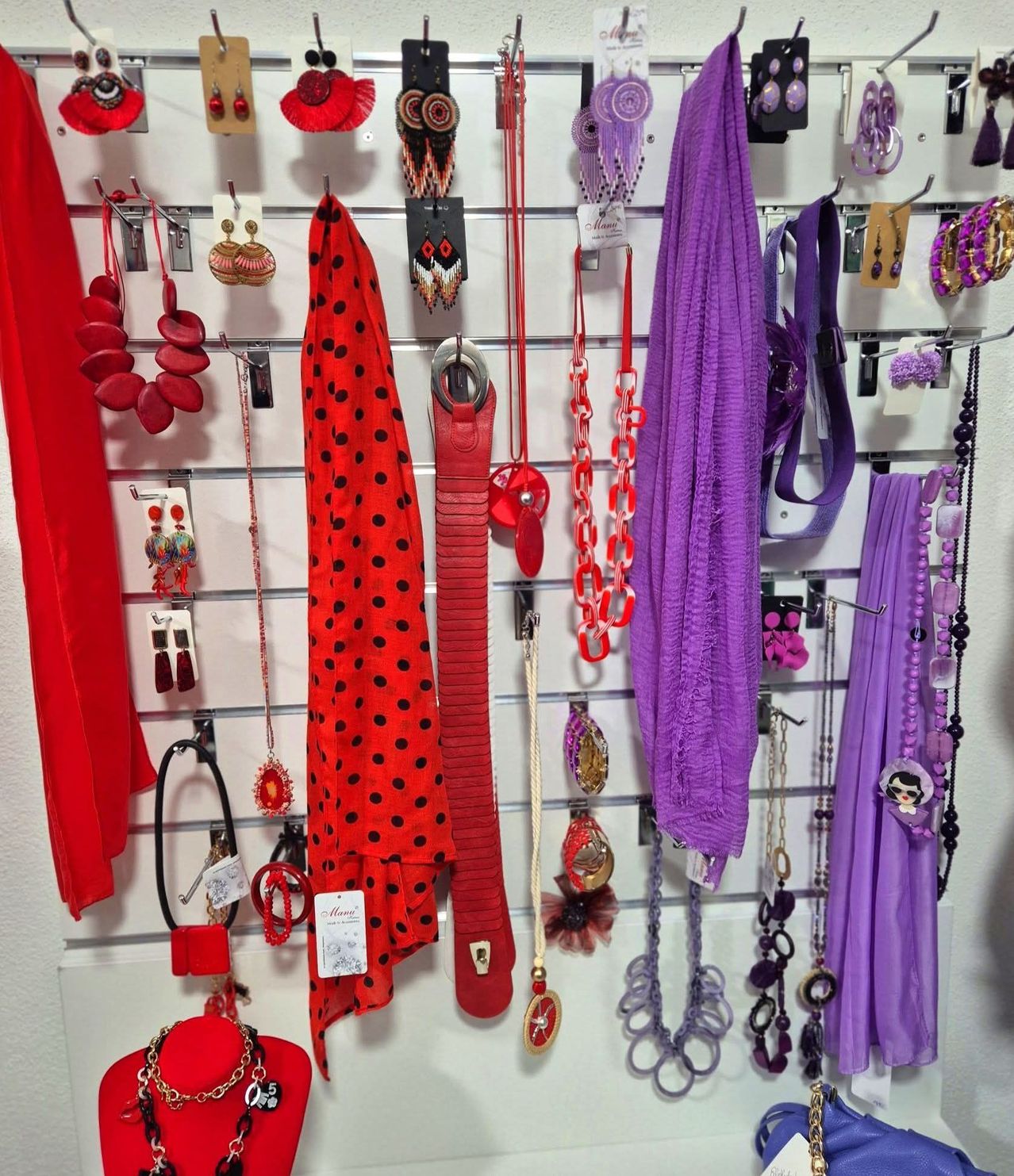 An einer weißen Wand sind verschiedene Accessoires in leuchtenden Farben wie Rot, Lila und Schwarz ausgestellt. Die Artikel sind ordentlich an Haken aufgereiht und zeigen eine Mischung aus Punkten, Ketten und komplizierten Designs.