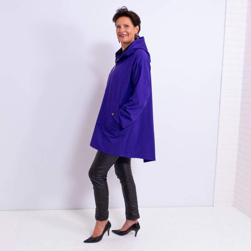 Eine Frau in einem lila Kapuzenmantel, schwarzen Lederhosen und hohen Absätzen posiert vor einem weißen Hintergrund.