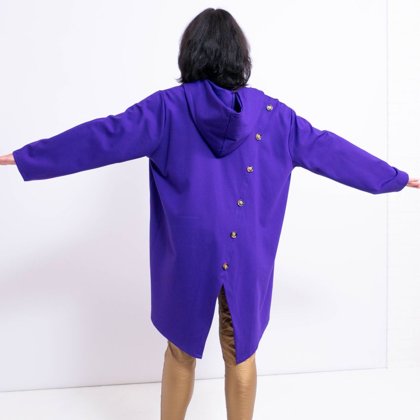 Eine Frau steht mit ausgestreckten Armen und trägt einen lila Kapuzenmantel mit goldenen Knöpfen und goldene Hosen vor weißem Hintergrund.