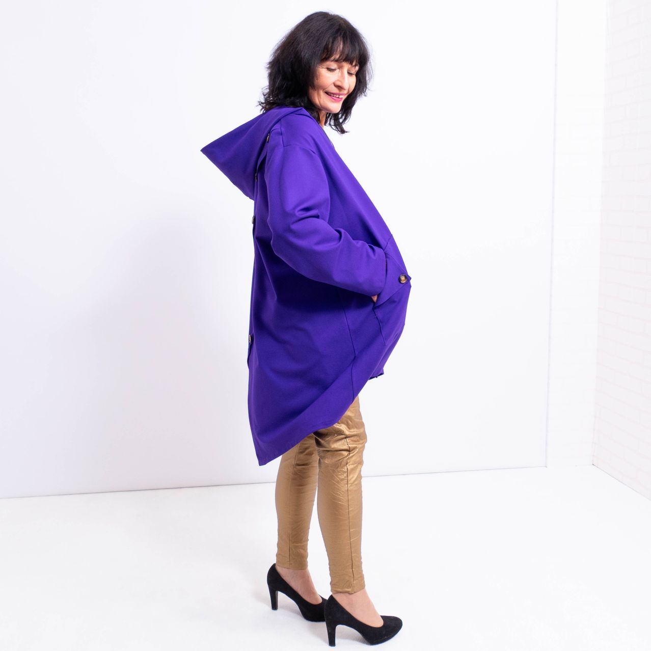 Eine Frau in einem lila Mantel mit Kapuze und braunen Hosen steht in einem weißen Raum und lächelt.