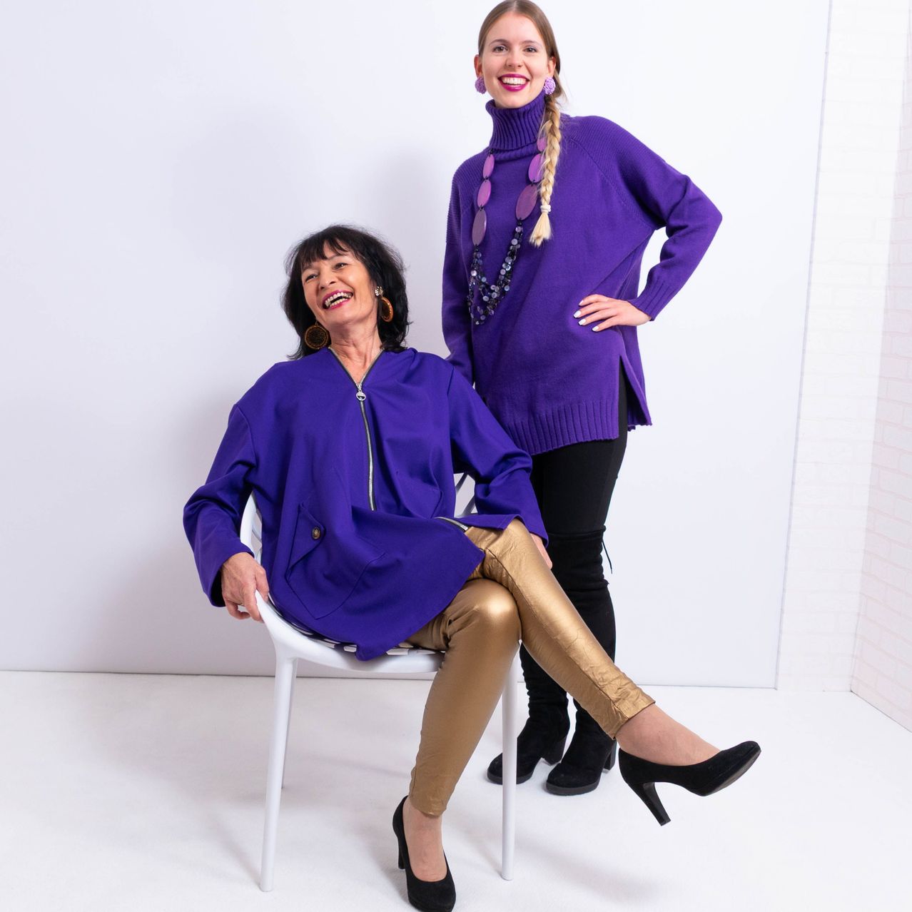 Zwei Frauen, eine auf einem Stuhl sitzend und die andere hinter ihr stehend, beide in lila Outfits, lächelnd und posieren für ein Foto.