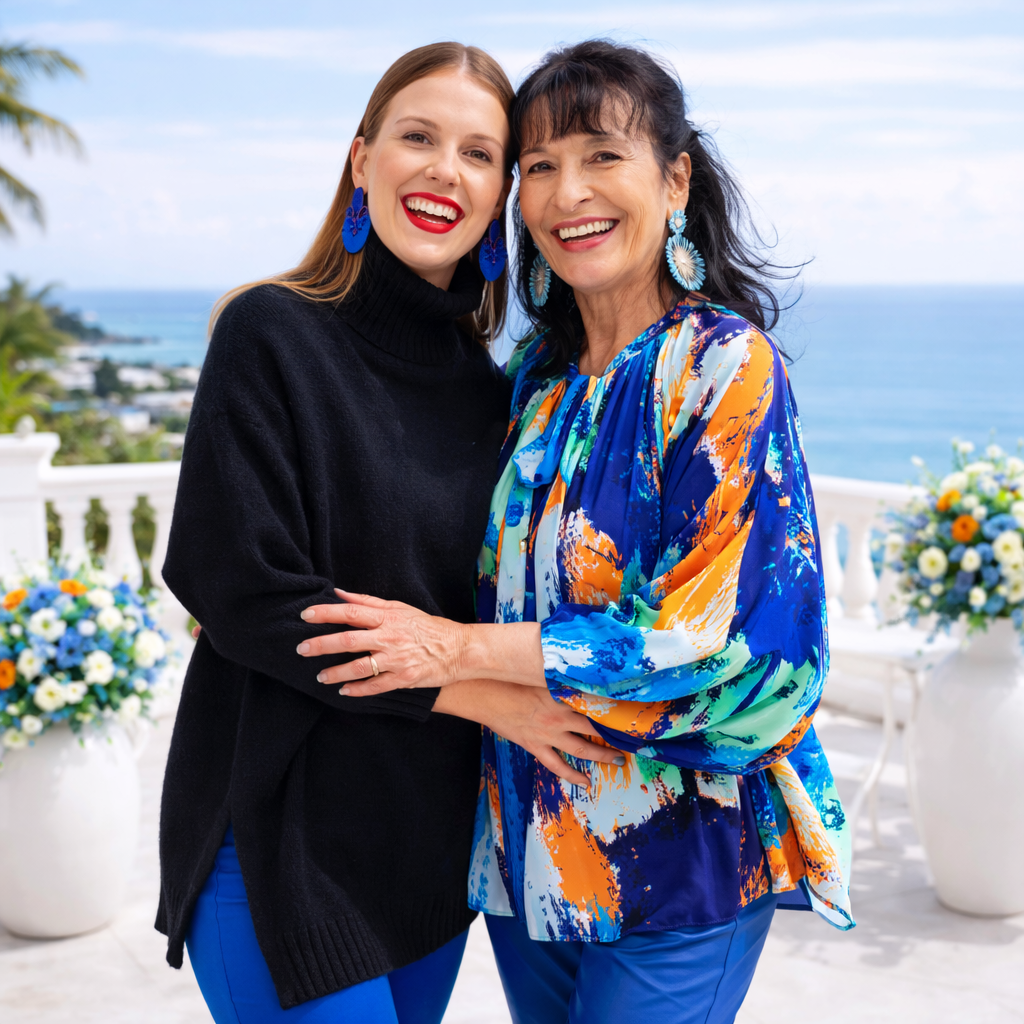 Zwei Frauen, eine in einem schwarzen Rollkragenpullover und die andere in einer farbenfrohen Bluse, stehen lächelnd auf einem Balkon mit Meerblick, Blumen in Vasen hinter ihnen.
