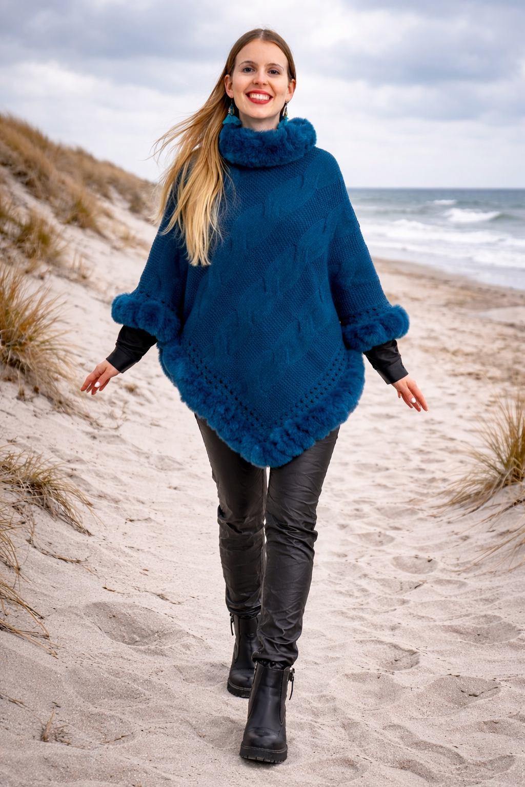 A woman in a blue poncho and black pants walks along a sandy beach near the ocean.