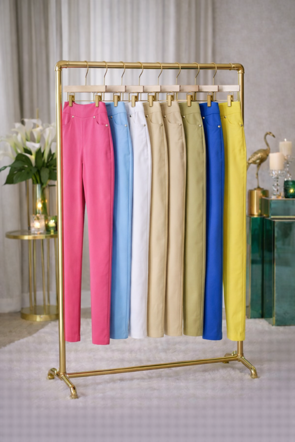 Ein Kleiderständer mit verschiedenen farbigen Hosen, darunter pink, blau, weiß, beige und gelb, in einem Raum mit weißen Vorhängen und einer Pflanze.