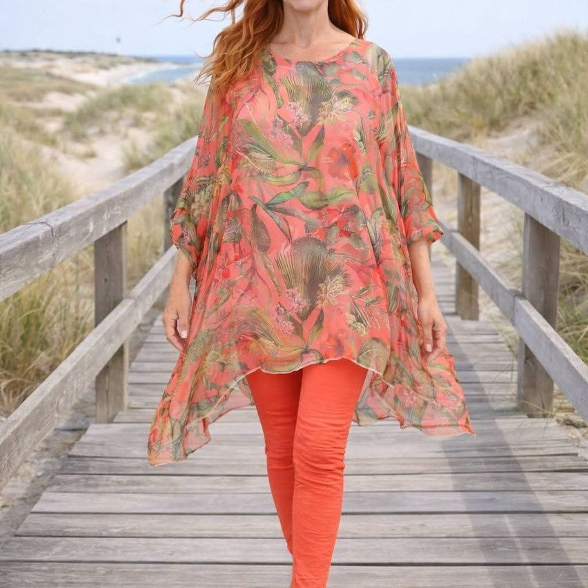 Eine Frau mit roten Haaren geht auf einem Holzweg am Strand, trägt eine korallenfarbene Bluse mit Blumenmuster und orangefarbene Hose.