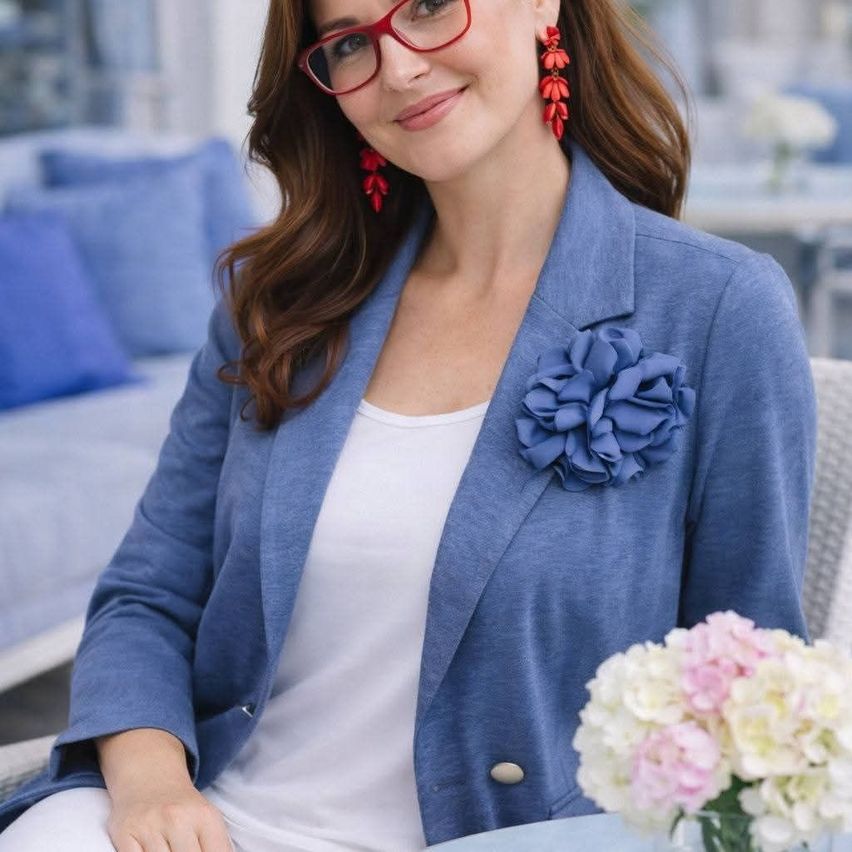 A woman with red earrings and a blue blazer with a blue flower pin sits on a chair with a table of flowers in front of her.