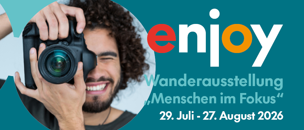 Ein lächelnder Mann mit lockigem Haar hält eine Kamera mit den Worten enjoy Wanderausstellung Menschen im Fokus und den Daten 29. Juli - 27. August 20.