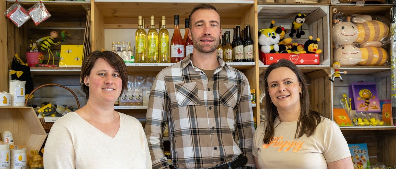 Drei Personen stehen in einem Raum voller Holzregalen, die verschiedene Produkte wie Flaschen, Spielzeug und Kisten präsentieren. Ein Mann in einem karierten Hemd steht in der Mitte, während zwei Frauen auf beiden Seiten in die Kamera lächeln.