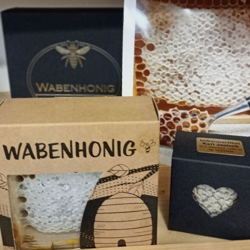 Verschiedene Schachteln Wabenhall-Honig mit einer Biene auf der Vorderseite, auf einer Holzoberfläche platziert. Ein herzförmiges Kästchen befindet sich rechts, mit einem goldenen Etikett.