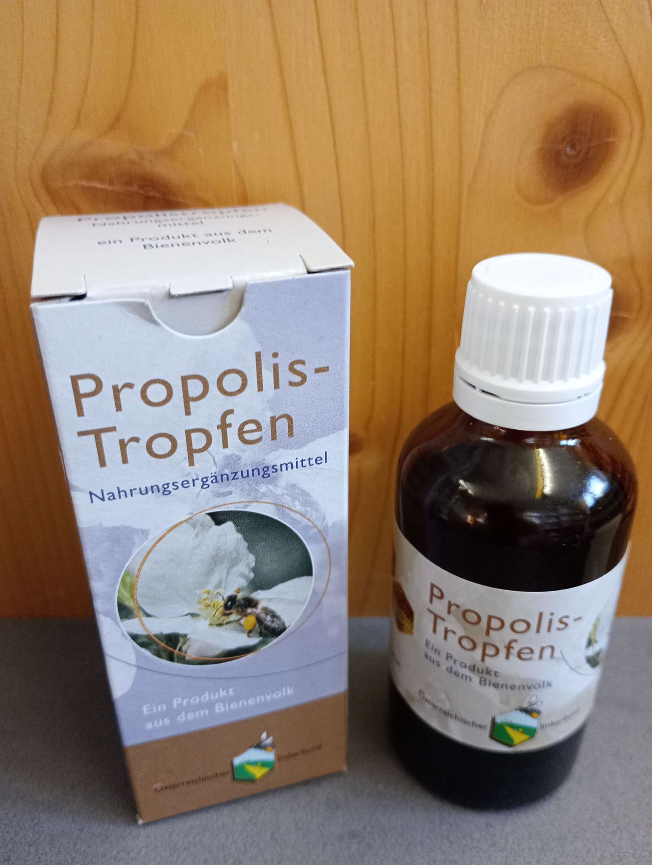 Eine Flasche und eine Schachtel Propolis Tropfen stehen auf einem holzfarbenen Hintergrund. Die Flasche ist voll und hat einen weißen Deckel. Die Schachtel hat ein Bild einer Biene auf einer Blume.