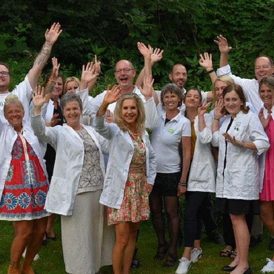 Eine Gruppe von Menschen in weißen Kitteln steht lächelnd in einem Garten und winkt mit den Händen.