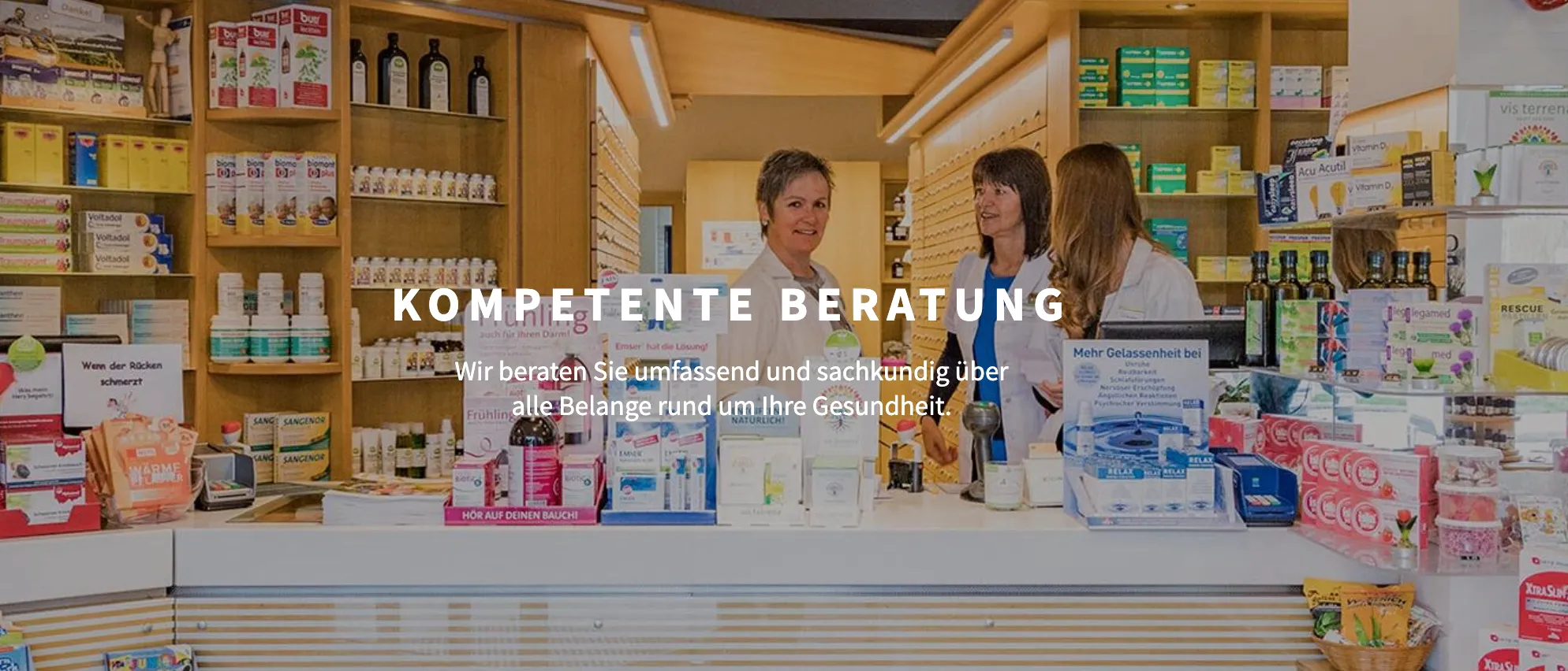 Drei Frauen in weißen Laborkittel stehen hinter einer Apothekentheke, lächeln und führen ein Gespräch. Die Theke ist mit verschiedenen Gesundheitsprodukten und Nahrungsergänzungsmitteln bestückt. Über ihnen steht in deutscher Schrift 'MPETENTE BERATUNG'.