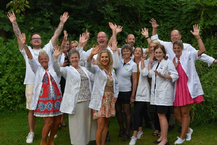 Eine Gruppe von Menschen in weißen Kitteln und Freizeitkleidung posiert für ein Foto und hebt die Hände, vor einem Hintergrund von Grün.