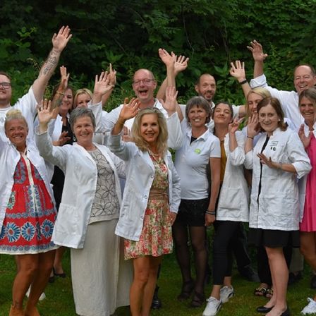 Eine Gruppe von Menschen in weißen Kitteln und Freizeitkleidung posiert für ein Foto und hebt die Hände, vor einem Hintergrund von Grün.