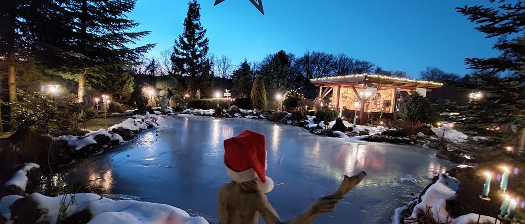 Ein gefrorener Teich mit einer Statue eines Mannes mit einem Weihnachtshut, umgeben von Weihnachtsdekoration und einem Haus mit Lichtern. Bäume und Büsche sind im Hintergrund.