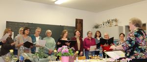 Eine Gruppe von Frauen mit Brillen und Mänteln versammelt sich in einem Raum, hält Bücher und Blumen, mit einem Tisch und Trophäen im Hintergrund.