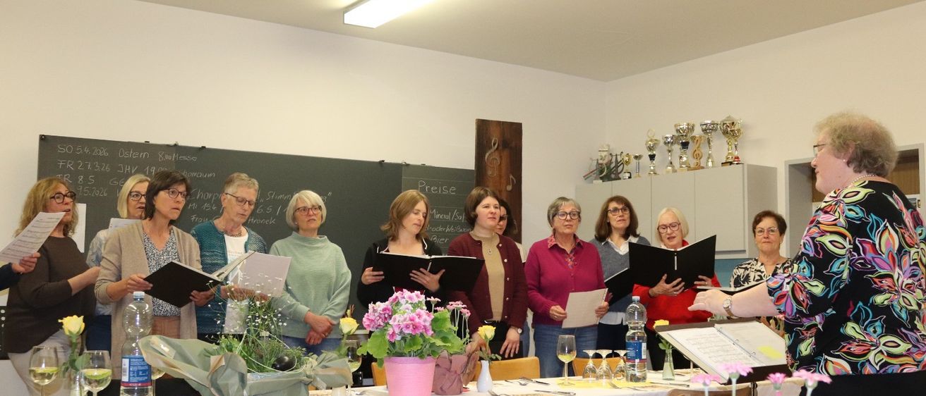 Eine Gruppe von Frauen mit Brillen und Mänteln versammelt sich in einem Raum, hält Bücher und Blumen, mit einem Tisch und Trophäen im Hintergrund.