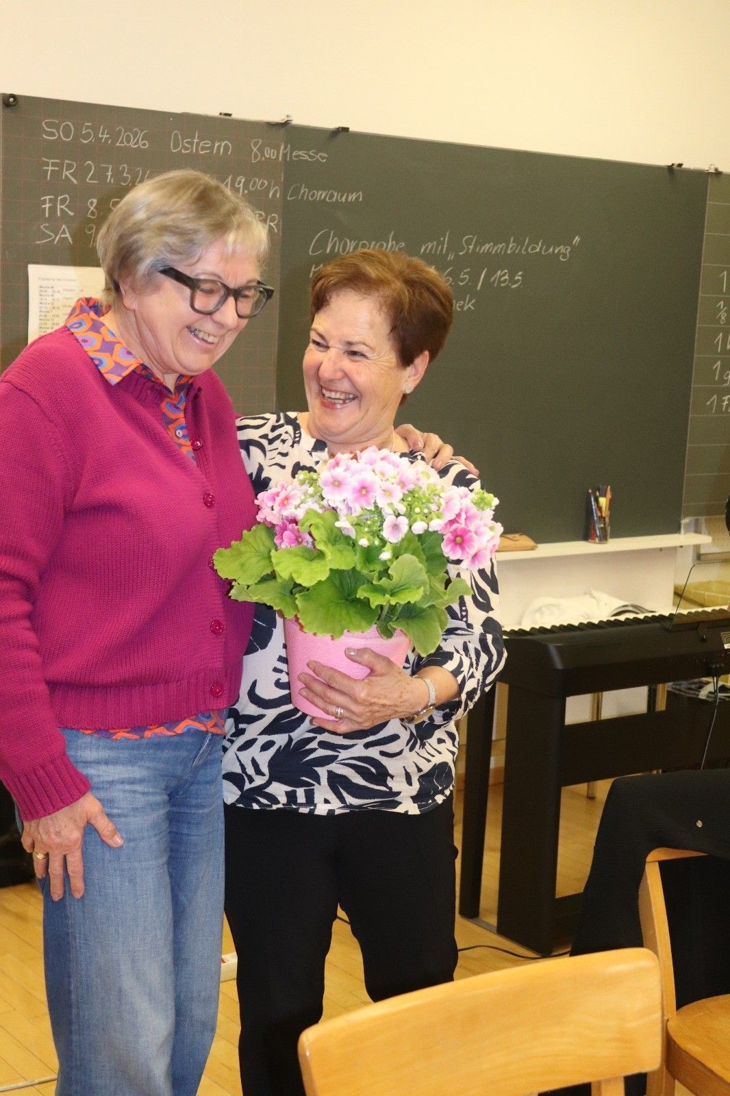 Zwei Frauen, beide mit Brille, stehen eng beieinander mit einem Lächeln auf den Gesichtern. Die Frau auf der rechten Seite hält einen pinkfarbenen Blumentopf.